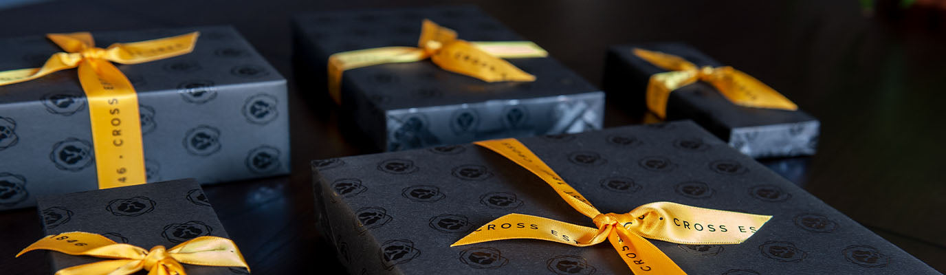 Assorted gifts wrapped in Cross-branded wrapping paper with yellow ribbons, arranged together.