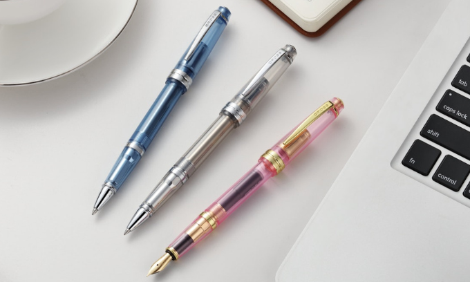 Bailey Light Translucent pens displayed on a desk, including the Translucent Navy Blue ballpoint pen, the Translucent Clear ballpoint pen, and the Translucent Sapphire Pink fountain pen.